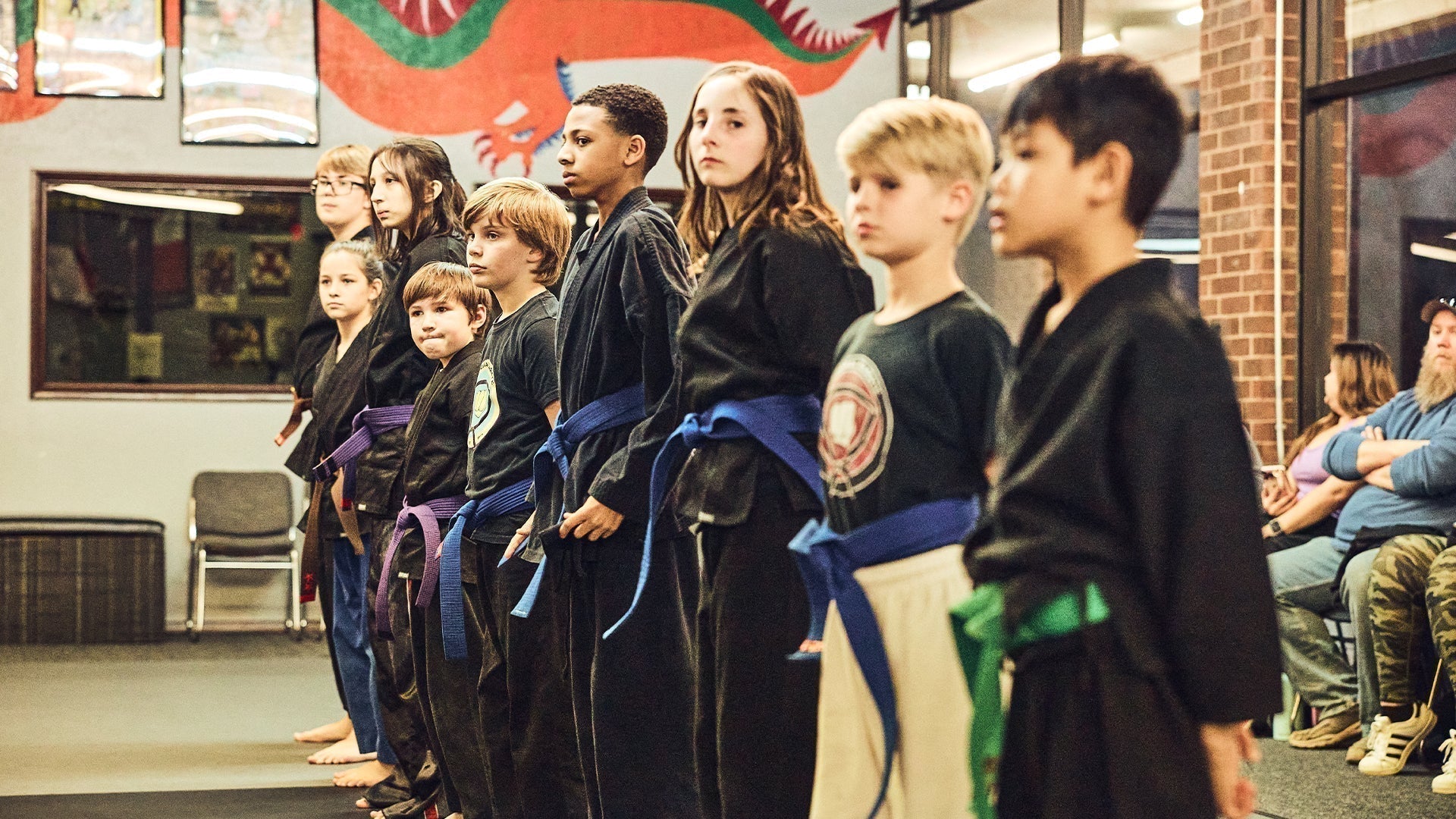 Children of different ages in black martial arts uniforms stand in a line with colored belts during a youth class or belt testing ceremony at a dojo.